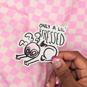 A sticker featuring a wide eyed and visibly exhausted cartoon character slouched over themselves on the floor, with text above the illustration reading, “only a lil’ stressed.’ The sticker is being held up in someone’s hand.