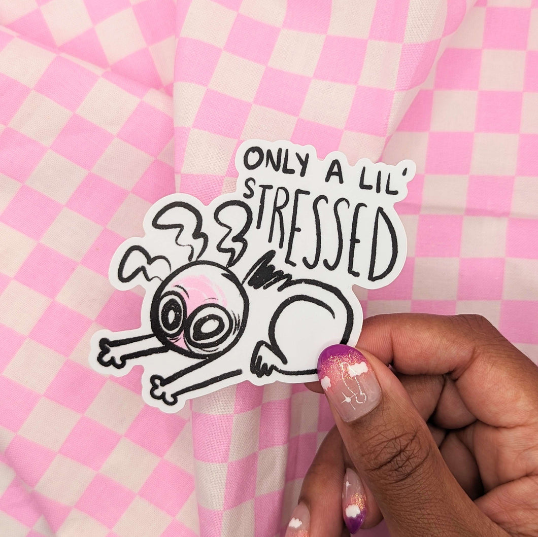 A sticker featuring a wide eyed and visibly exhausted cartoon character slouched over themselves on the floor, with text above the illustration reading, “only a lil’ stressed.’ The sticker is being held up in someone’s hand.