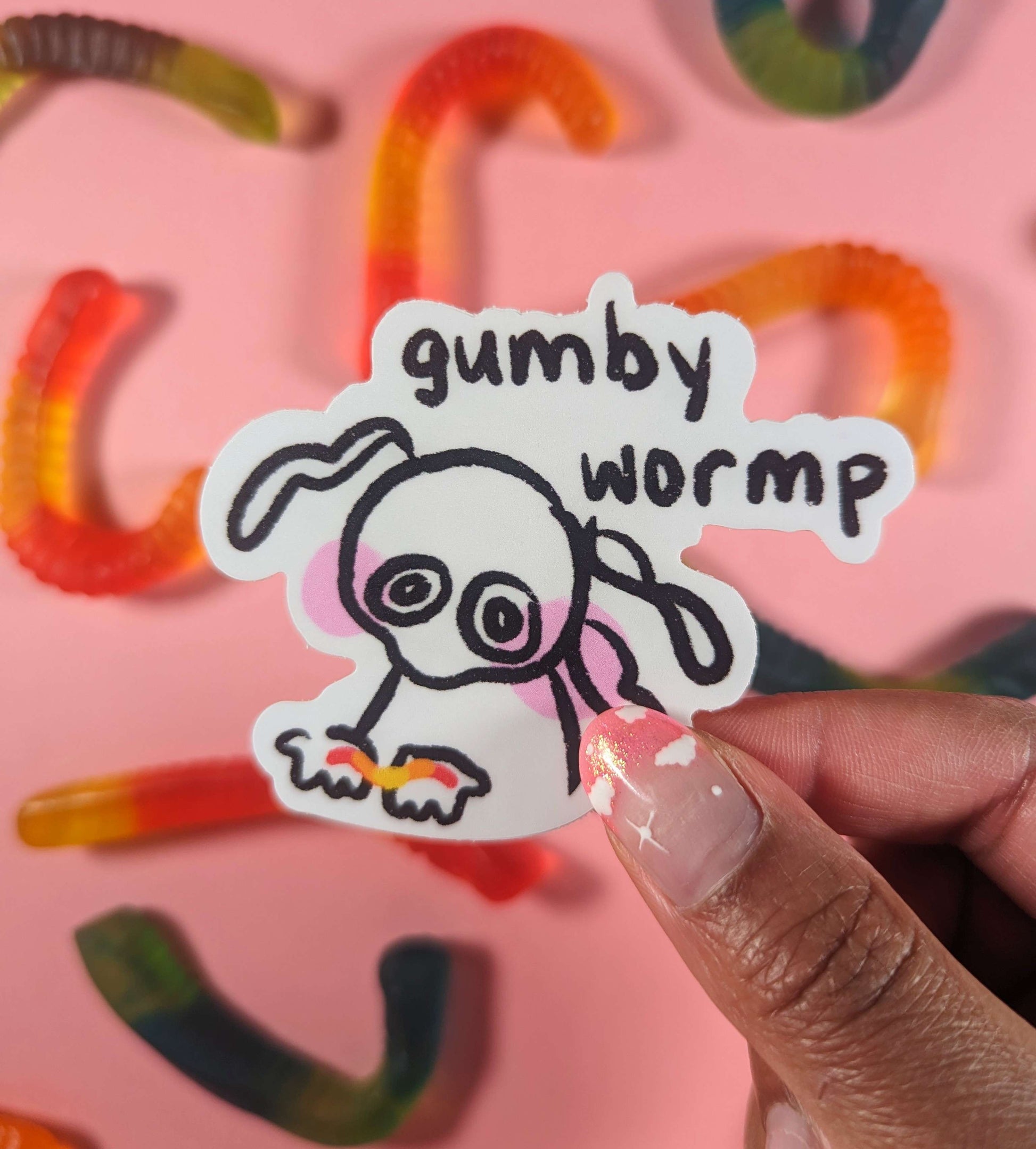 A sticker featuring a cute character gently holding a colorful gummy worm in the palms of their hands. Text above it reads, 'gumby wormp.' The sticker is held in front of a pink gummy worm patterned background.