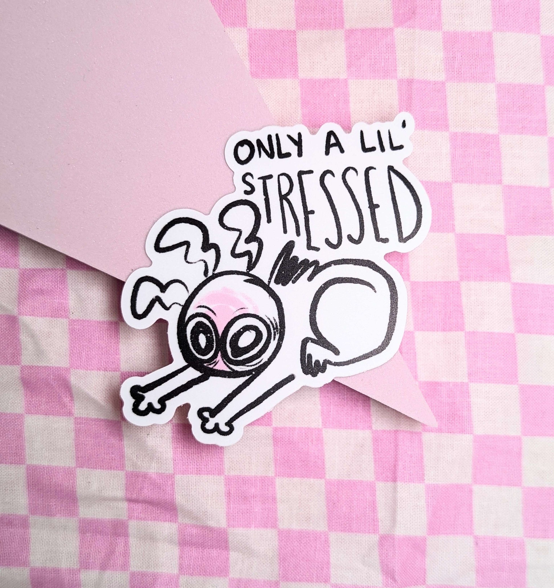 A sticker featuring a wide eyed and visibly exhausted cartoon character slouched over themselves on the floor, with text above the illustration reading, “only a lil’ stressed.’ The sticker is laying on a pink checkered background.