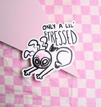 Load image into Gallery viewer, A sticker featuring a wide eyed and visibly exhausted cartoon character slouched over themselves on the floor, with text above the illustration reading, “only a lil’ stressed.’ The sticker is laying on a pink checkered background.
