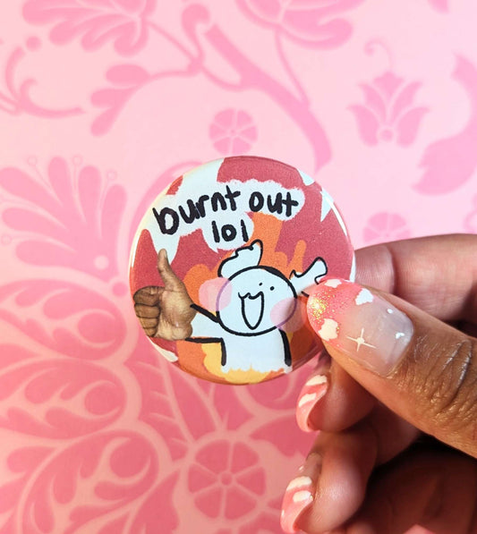 A round button pin featuring a simple illustration of a character holding up a jarringly photoshopped hyperrealistic thumbs up. The character’s expression is cheerful, and they are surrounded by cartoon flames. Text above them reads, ‘burnt out lol.’